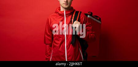 Consegna ragazzo dipendente uomo in rosso uniforme lavoro come corriere concessionario tenere rosso sacchetto termico cibo zaino isolato su sfondo rosso studio Foto Stock