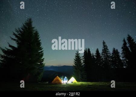 Luminose tende turistiche illuminate che brillano sul campeggio in montagne scure sotto il cielo notturno con stelle scintillanti. Concetto di stile di vita attivo Foto Stock