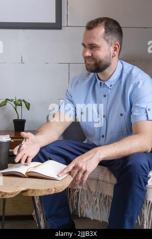 Un uomo arabo di mezza età in una tuta da lavoro che parla a un tavolo da caffè. Uomo d'affari sorridente che scrive le note nel diario personale, giorno di lavoro di pianificazione Foto Stock