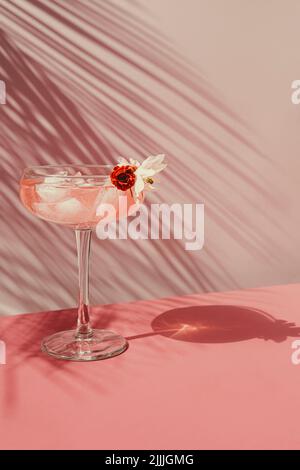 Cocktail estivo con champagne rosa con ghiaccio al tavolo con ombre dal sole. Sullo sfondo di una parete rosa con foglie di palma, vista frontale. Foto Stock