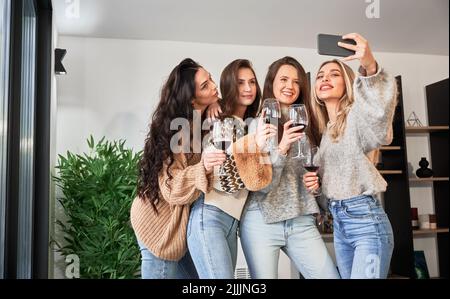 Le giovani donne che si divertiscono nei fine settimana invernali all'interno di una caserma contemporanea. Quattro ragazze che si divertono, prendono selfie al telefono e bevono vino. Foto Stock