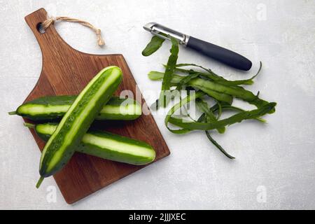 Cetrioli freschi con pelle e coltello su un tagliere di legno marrone su un tavolo di cemento grigio chiaro Foto Stock