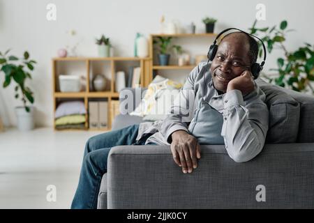 Ritratto di un anziano africano in cuffie sorridenti alla fotocamera mentre si riposa sul divano in salotto e si gode la musica Foto Stock
