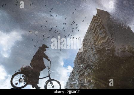 Sagoma sfocata di riflessione della persona su una bicicletta che guida da sola sul marciapiede bagnato della città nelle giornate piovose. Gli uccelli volano attraverso il cielo. Foto astratta Foto Stock