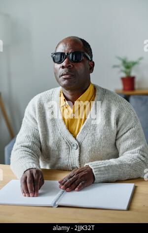 Uomo anziano cieco in occhiali scuri usando le mani per leggere in braille seduto al tavolo nella stanza Foto Stock