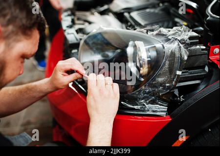 Addetto al servizio di assistenza auto ha messo una pellicola anti-ghiaia su una carrozzeria rossa presso l'officina del veicolo di dettaglio. Protezione dell'auto con pellicole speciali. Foto Stock