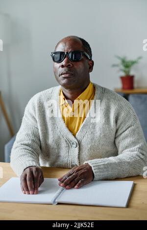 Uomo anziano cieco in occhiali scuri usando le mani per leggere in braille seduto al tavolo nella stanza Foto Stock