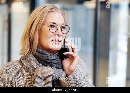 Donna d'affari matura in occhiali e in cappotto caldo sorridente durante la conversazione sul telefono cellulare Foto Stock