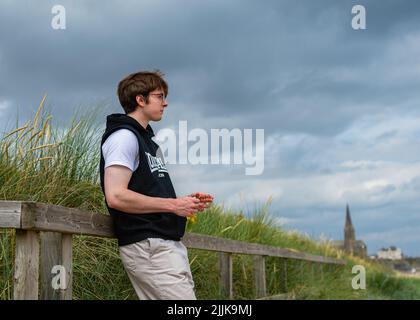 Ritratto naturale di un giovane uomo in piedi su una recinzione sul lungomare di LongSands spiaggia a Tynemouth con la chiesa sullo sfondo Foto Stock