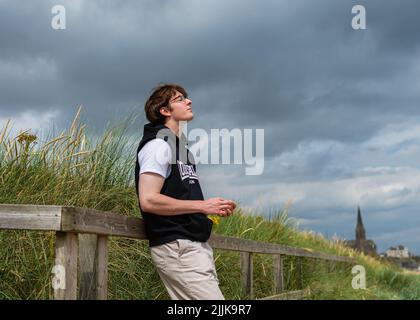 Ritratto naturale di un giovane uomo in piedi su una recinzione sul lungomare di LongSands spiaggia a Tynemouth con la chiesa sullo sfondo Foto Stock