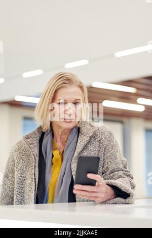 Donna d'affari matura in cappotto caldo che legge un messaggio sul telefono cellulare mentre si trova vicino alla reception nella sala degli uffici Foto Stock