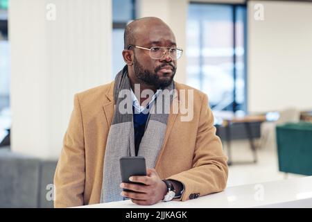Uomo d'affari africano in elegante cappotto utilizzando il suo telefono cellulare mentre si trova nella lobby vicino alla reception Foto Stock