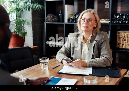 Donna d'affari matura in occhiali seduti al tavolo con documenti e utilizzando il suo telefono cellulare durante l'incontro con la collega al ristorante Foto Stock