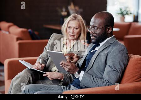 L'uomo d'affari africano che punta allo schermo del pc del tablet con la donna d'affari che fa le note nel taccuino, progettano il lavoro insieme seduto nel caffè Foto Stock