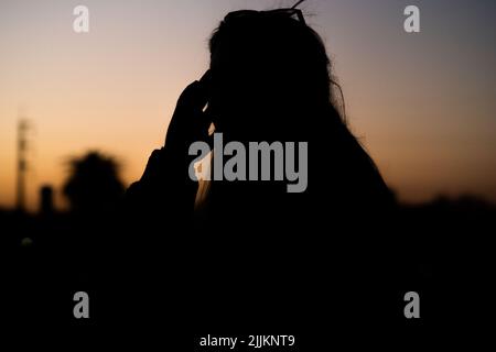 Fuoco selettivo della silhouette di una ragazza con capelli sciolti che tengono la testa con il tramonto sullo sfondo Foto Stock