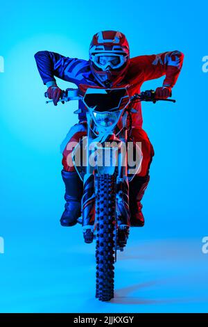 Ritratto di giovane uomo concentrato, biker che sviluppa velocità su moto isolato su sfondo blu studio in neon luce Foto Stock