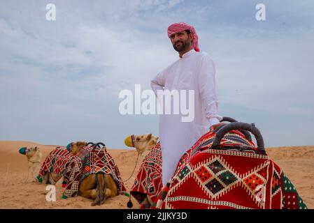 Un maschio arabo in twabh in piedi vicino ai suoi cammelli su un deserto Foto Stock