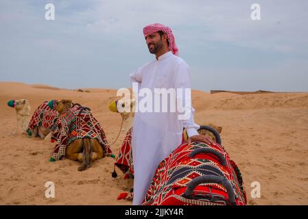 Un maschio arabo in twabh in piedi vicino ai suoi cammelli su un deserto Foto Stock