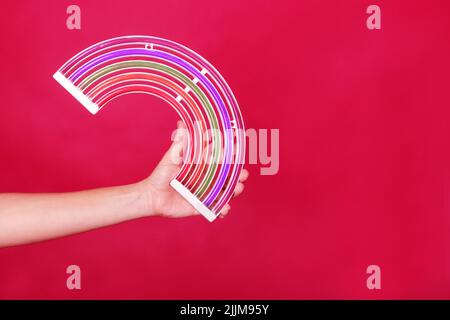 mano di donna caucasica che tiene un arcobaleno al neon su un rosso background.jpg Foto Stock