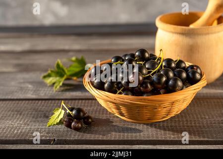 Ribes nero maturo in una ciotola di vimini e un mortaio per fare la marmellata su un tavolo di legno al tramonto con ramoscelli e foglie. Foto Stock