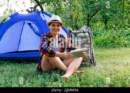 Defocus giovane donna che lavora sul tablet vicino campeggio tenda all'aperto circondato da bella natura. Freelance, sabbatico, salute mentale. Persona online. Foto Stock