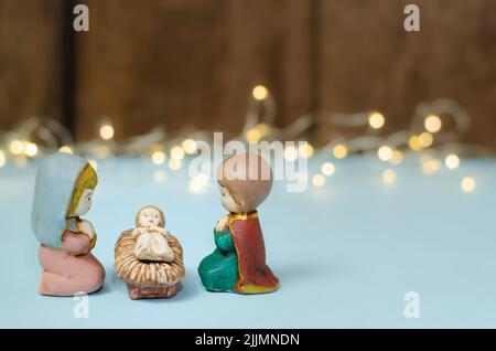 A close-up shot of mini colorful figures of the Nativity scene on a white table with string of lights and brown wooden wall in the blurred background Stock Photo