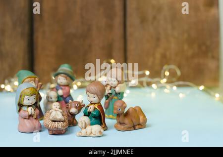 Un primo piano di mini figure colorate della scena Natività su un tavolo bianco con una serie di luci e una parete di legno marrone sullo sfondo sfocato Foto Stock