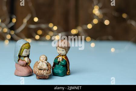 Un primo piano di mini figure colorate della scena Natività su un tavolo bianco con una serie di luci e una parete di legno marrone sullo sfondo sfocato Foto Stock