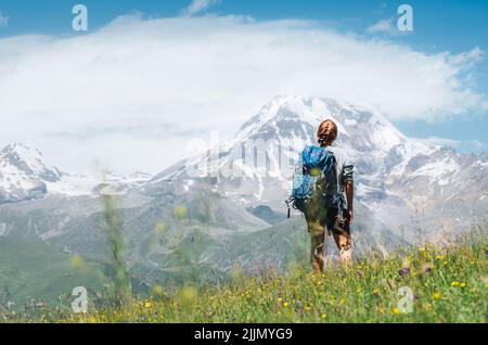 Donna Backpacker con uno zaino in piedi e godere di piste innevate di montagna Kazbek 5054m mentre lei cammina da verde erba collina. Moun del Caucaso orientale Foto Stock