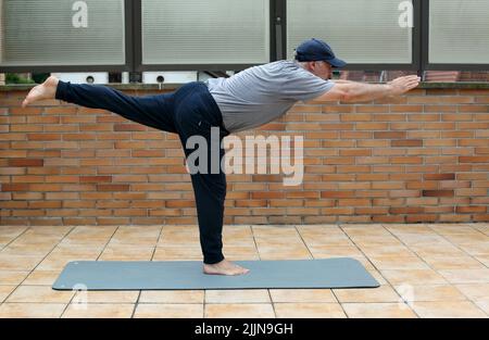 Una vista di un uomo caucasico maturo in abbigliamento sportivo che fa esercizio di yoga (bilanciamento della posa del bastone) durante il giorno Foto Stock