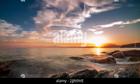 Una lunga esposizione di un mare che lava la spiaggia rocciosa al tramonto nuvoloso Foto Stock