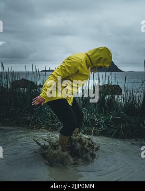 colpo verticale di ragazza in impermeabile giallo che salta in pozzanghera contro di oceano Foto Stock