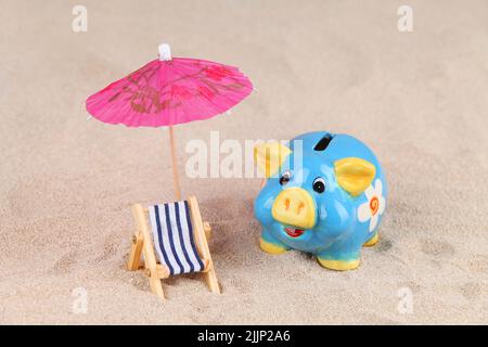 Una banca di piggy blu e giallo, ombrello e lettino sulla sabbia - risparmiare denaro per le vacanze Foto Stock