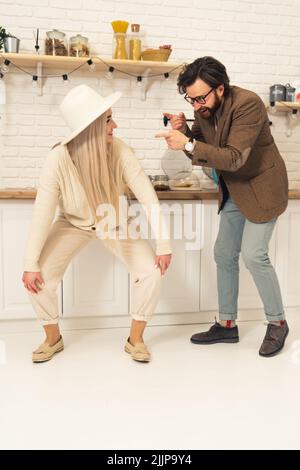 Giovane coppia caucasica che fa danze divertenti insieme in cucina. Donna bionda che indossa un cappello e un uomo dai capelli scuri con barba. Scatto verticale per interni. Foto di alta qualità Foto Stock