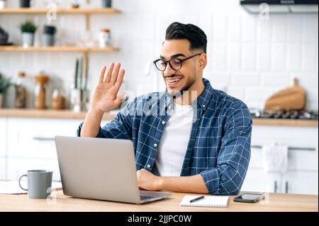 Amichevole felice arabo o indiano uomo con occhiali, seduto a casa in cucina, conduce videochiamata con il cliente, incontro virtuale con il collega, utilizza il laptop, fare gesto di saluto, sorridente Foto Stock