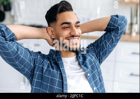 Primo piano di un ragazzo arabo o indiano felice e rilassato, freelance, che lavora da casa, si siede in cucina, prende una pausa dal lavoro, mette le mani dietro la testa, guarda direttamente, sognando, sorride Foto Stock