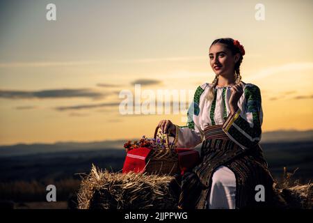 Una giovane donna in un abito tradizionale rumeno con un cesto di fiori su un campo al tramonto Foto Stock