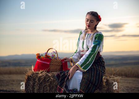 Una giovane donna in un abito tradizionale rumeno con un cesto di fiori su un campo al tramonto Foto Stock
