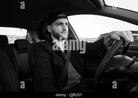 Un'immagine in scala di grigi di un bell'uomo caucasico che guida un'auto che indossa una cintura di sicurezza Foto Stock