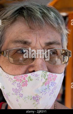Un ritratto di una donna ispanica anziana con occhiali che indossano una maschera facciale riutilizzabile in tessuto all'aperto in una giornata di sole Foto Stock
