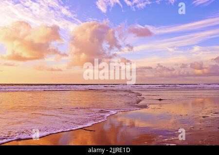 Bella spiaggia e fresco oceano contro un cielo nuvoloso morbido al tramonto con spazio copia. Onde che si lavano su una riva su una costa rilassante e panoramica. Dorato Foto Stock