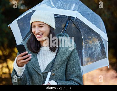 Controllare se sta per piovere di nuovo. Una giovane donna attraente in piedi da sola fuori e usando il suo cellulare mentre tiene un ombrello. Foto Stock