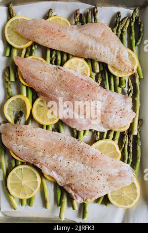 Pesce rockfish pacifico crudo su un letto di asparagi e ruote di limone, foglio piatto cena. Fare la trota Amandine. Foto Stock