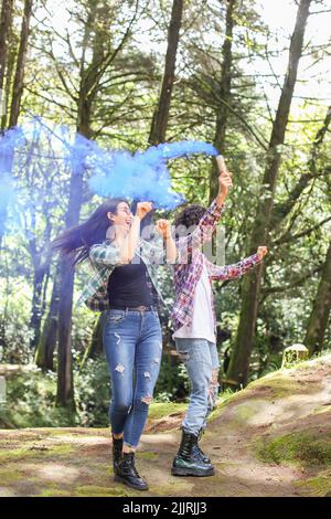 Giovane coppia latino-americana che balla nella foresta sorridendo tenendo una bomba di fumo di colore blu in Costa Rica Foto Stock