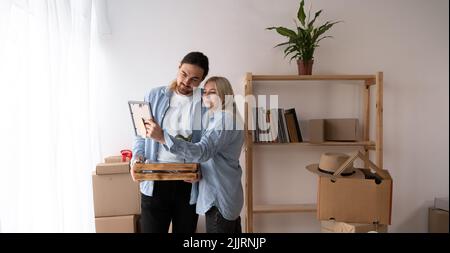 Giovane coppia felice che tiene la cornice ammira la pittura o la foto al giorno del trasferimento nella nuova prima casa. Concetto di ristrutturazione, nuovo negozio di mobili Foto Stock