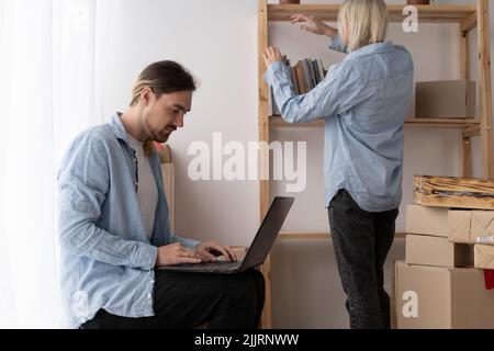Appena spostato in. Coppia nell'amore che controlla le idee dell'interno per nuovo appartamento usando il laptop, l'uomo con il pc, la signora che disimpacchettano le cose, spazio della copia. In movimento Foto Stock