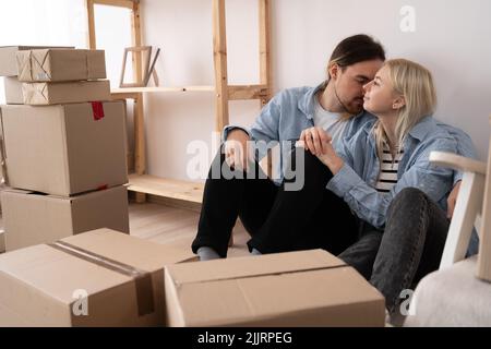 Felice giovane coppia seduta sul pavimento dell'appartamento appena affittato o acquistato. Scatole di cartone disimballate e mobili coperti nel moderno Foto Stock