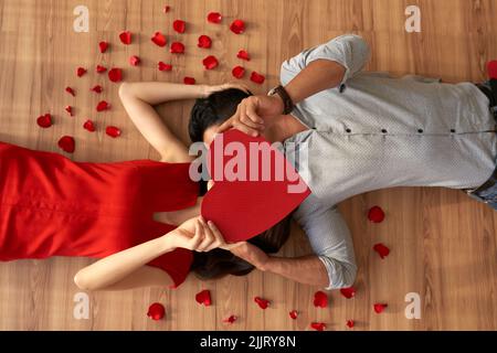 Direttamente sopra la vista di coppia giovane amorevole che copre i loro volti con pezzo di carta a forma di cuore e baciare mentre si trova sul pavimento, petali rossi di rosa sparsi ovunque Foto Stock