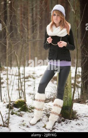 Un colpo verticale di una ragazza caucasica bionda che indossa un cappello bianco e una sciarpa in un parco innevato Foto Stock