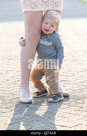 Triste ragazzo biondo caucasico che abbraccia le gambe della madre. Foto di alta qualità Foto Stock